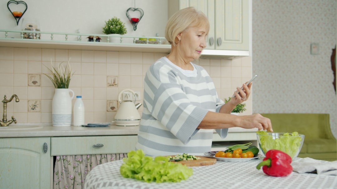 Old lady is cooking vegetable salad and looking at smartphone screen checking ingredients for meal during dinner preparation at home. People and gadgets concept.