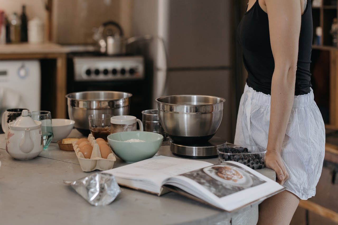 Woman in kitchen preparing breakfast with various ingredients and a recipe book open.